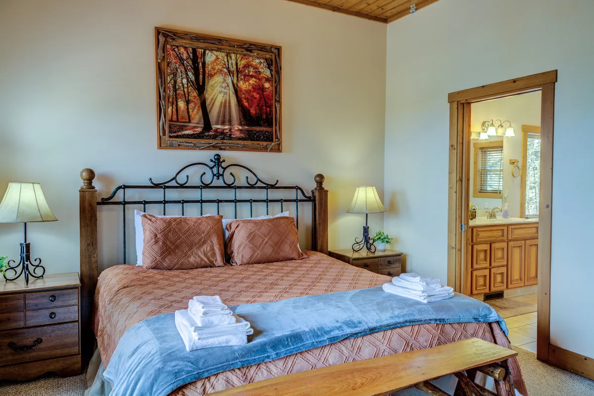 A tranquil bedroom featuring a plush bed adorned with decorative pillows, framed artwork on the wall, bedside tables with lamps, and an adjacent bathroom entrance. Soft, natural light filters through the space, enhancing the cozy and serene atmosphere.