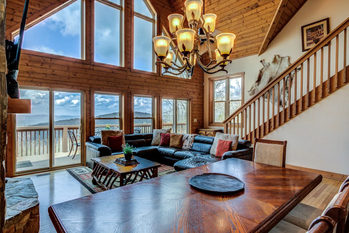 The image shows an inviting interior living space featuring a large sectional sofa arranged for gatherings, a striking chandelier, and expansive windows framing breathtaking long-range views of the Blue Ridge Mountains. A wooden staircase adds rustic charm, and the room is filled with natural light, creating a warm and welcoming atmosphere ideal for family or group relaxation.