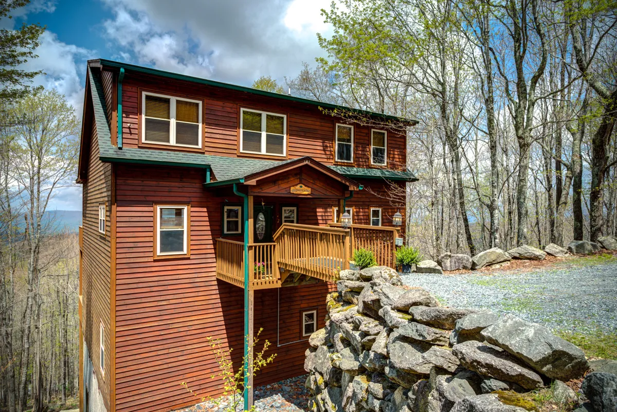 This exterior shot shows the wooden facade of the lodge, front porch, rock wall, and surrounding trees. The setting evokes a welcoming, tranquil atmosphere and hints at outdoor relaxation, family gatherings, and the possibility of evening firepit sessions.
