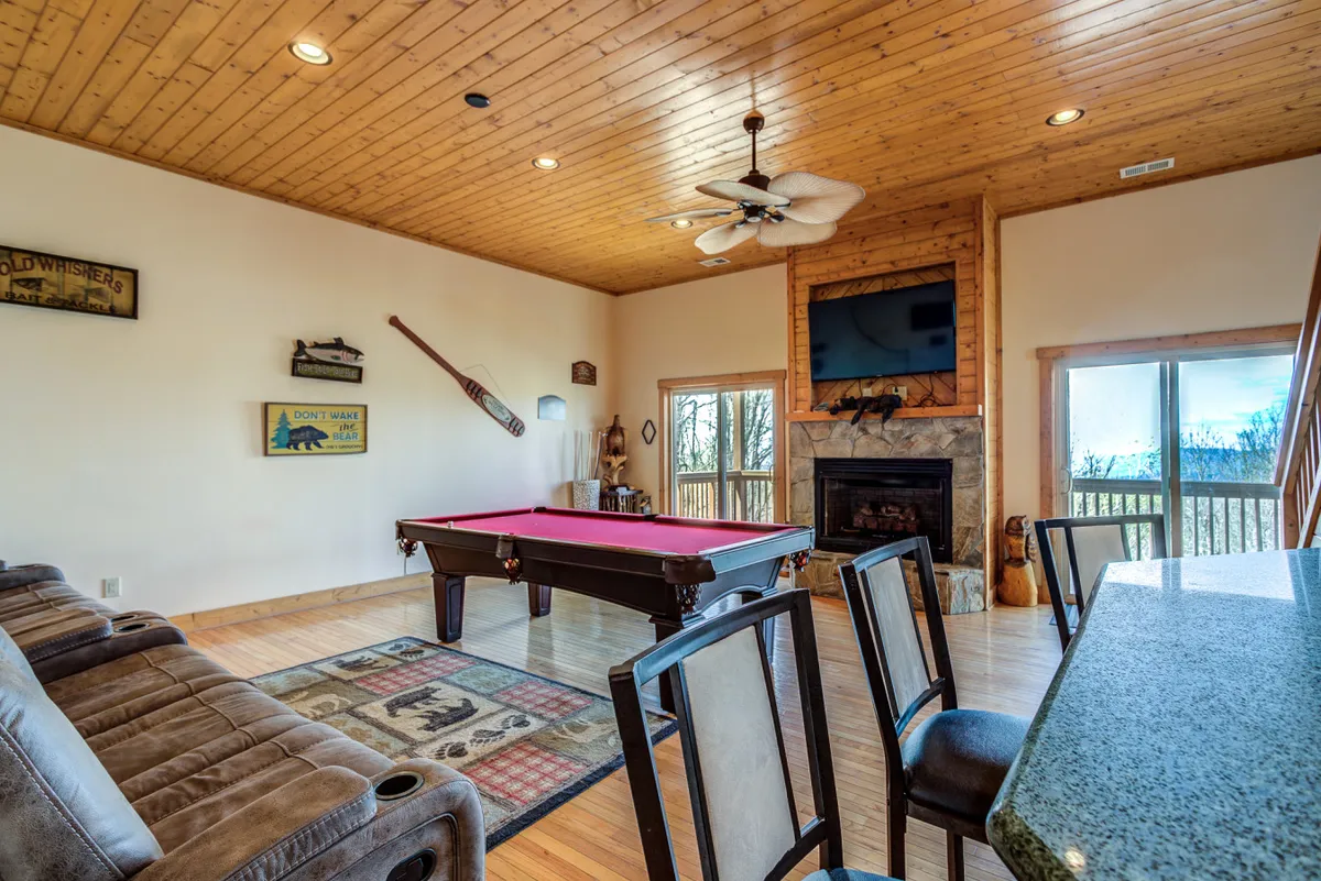 An interior entertainment area with a pool table, stone fireplace, large windows offering views, bar seating, and decorative touches. The setting is warm and lively, ideal for game nights or unwinding by the fire.