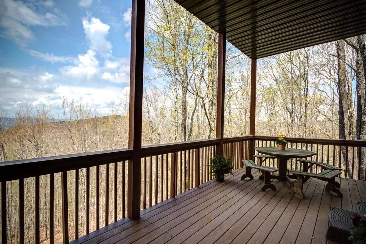 A wooden deck with a table and benches overlooks a peaceful natural landscape dotted with trees. The scene is tranquil, inviting guests to relax and enjoy the outdoors. The spacious deck offers ample room for family members, including pets, to spend time together while soaking in the fresh mountain air.