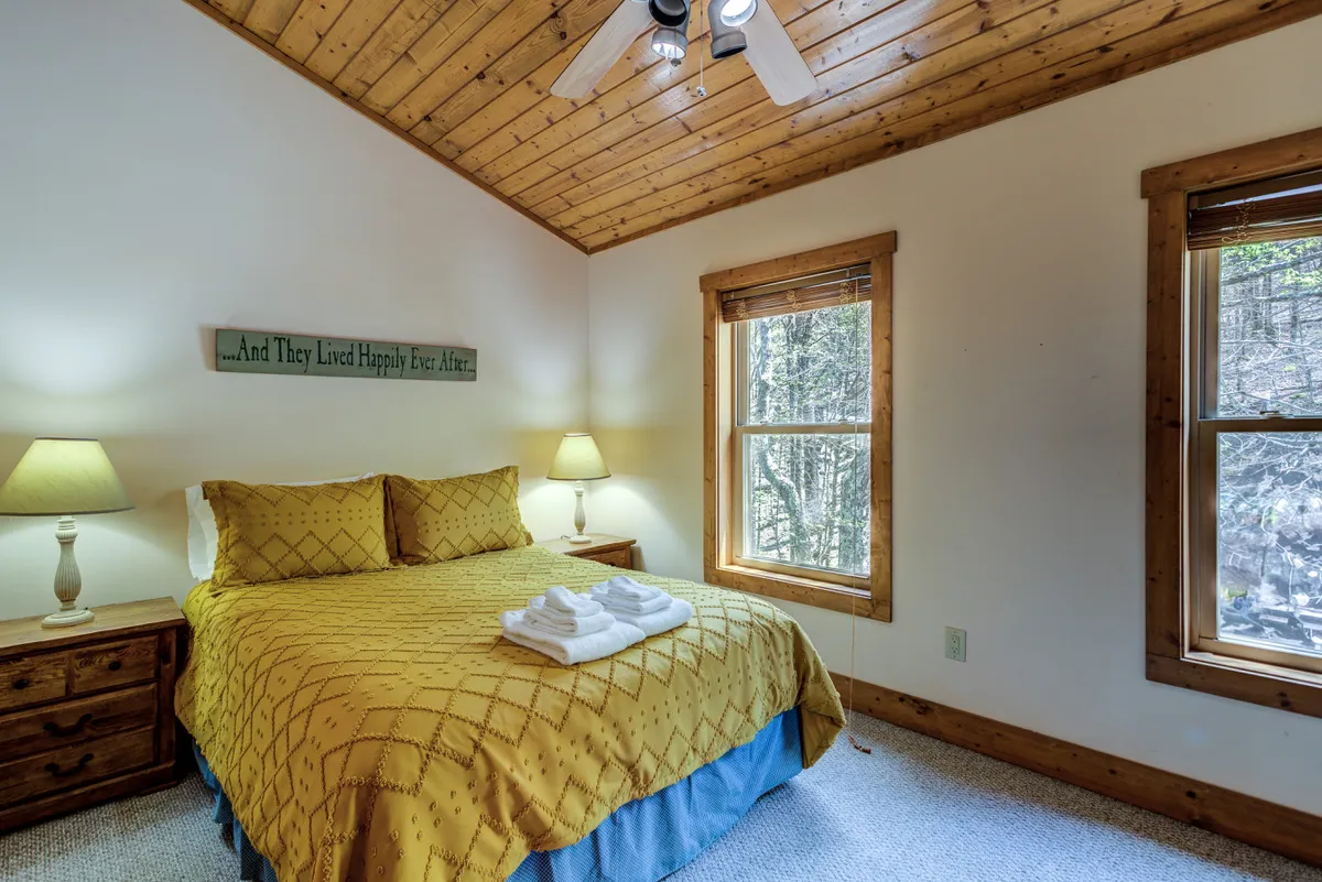 A serene bedroom with yellow bedding, twin bedside lamps, expansive windows offering views of the surrounding forest, and a warm wooden ceiling. The space is bathed in morning light, creating a peaceful retreat.
