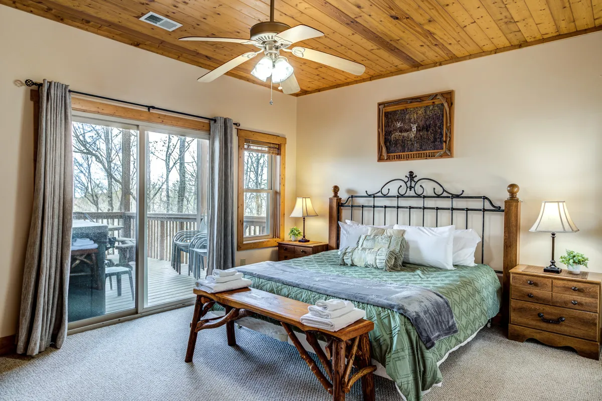 An inviting bedroom with a large bed dressed in decorative pillows, a wooden bench at the foot, expansive windows leading to a deck, and nightstands with lamps. Soft lighting and natural views create a restful atmosphere.