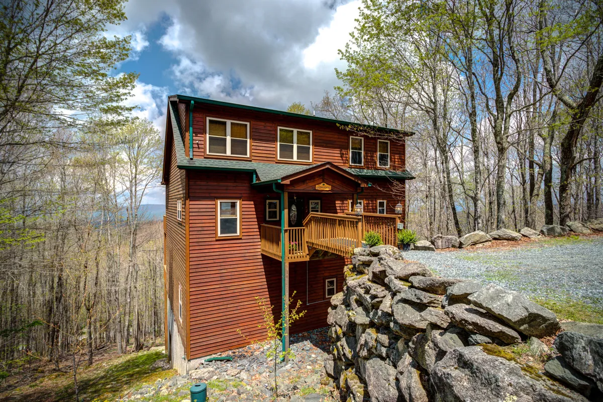 The wooden exterior of the house is surrounded by tall trees, with a balcony that invites guests to enjoy the outdoors. A rock wall in the foreground adds rustic charm, and the entire setting suggests proximity to wooded trails and natural exploration.