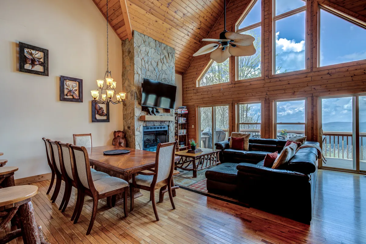 This interior image features a spacious living and dining area with large windows that frame scenic mountain views. The focal points include a stone fireplace, a comfortable seating arrangement, a wooden dining table, and warm, inviting decor. The natural light streaming in enhances the cozy and communal ambiance.
