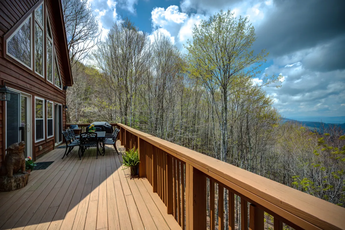 A spacious outdoor deck with an inviting dining set, surrounded by lush trees and offering expansive mountain views in the distance. The scene captures the potential for group meals, relaxation, and appreciation of the ever-changing foliage throughout the seasons.