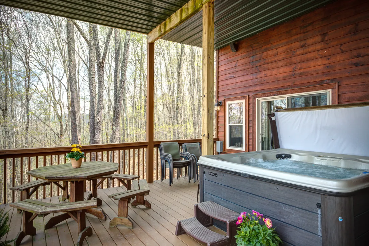 An expansive deck tucked among trees, with a six-person hot tub taking center stage. The scene includes a dining table and glimpses of surrounding nature, creating an inviting oasis for relaxation and outdoor enjoyment.