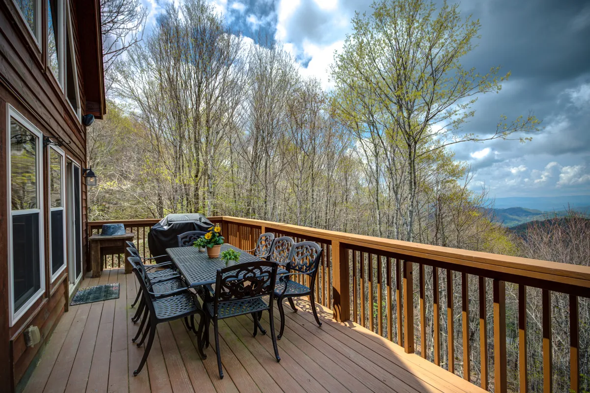 A serene deck setup with an outdoor dining table, a grill ready for barbecues, potted flowers, and a backdrop of dramatic mountain vistas. The table arrangement hints at group gatherings, outdoor meals, and lively evenings against a panoramic setting.